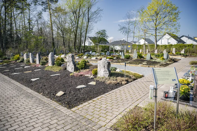 Blick über die Grabfelder des Erinnerungsgartens Schellbronn: dunkel gemulchte Beete mit aufgestellten Natursteinen, gepflasterte Wege und eine Informationstafel, dahinter Wohnhäuser am Ortsrand.