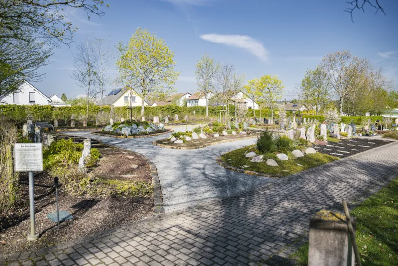 Übersicht über den Erinnerungsgarten mit stehenden Grabmalen, geschnittenen Hecken und gepflasterten Wegen, im Hintergrund Häuser des Ortes.